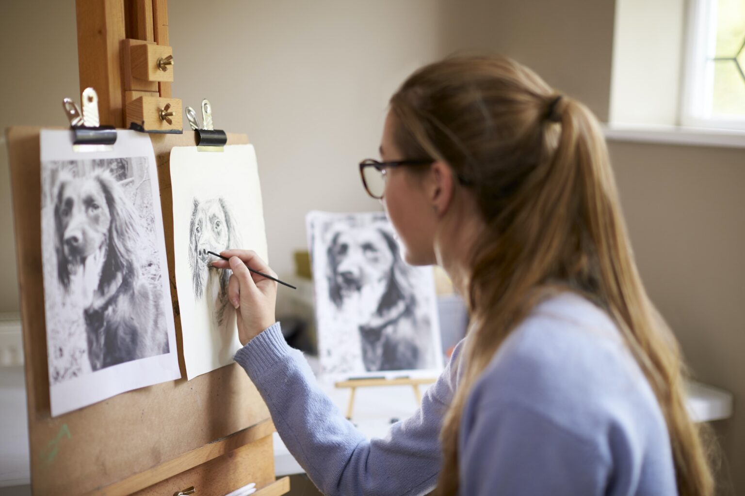 Side View Of Female Teenage Artist Sitting At Easel Drawing Picture Of Dog In Charcoal