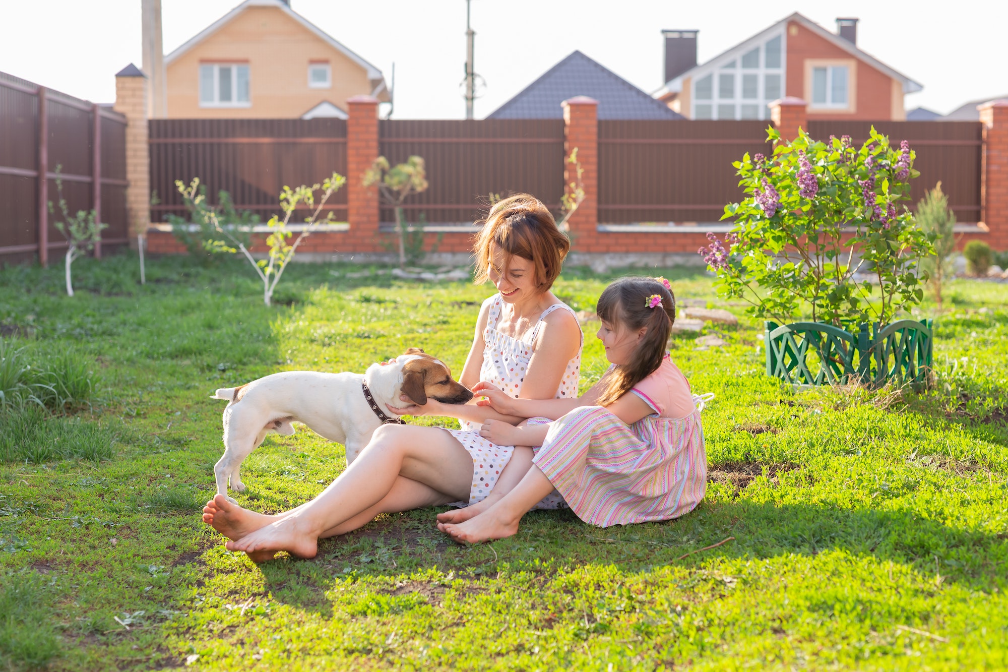 Cute young mother and daughter sit on the grass of their country house barefoot next to their