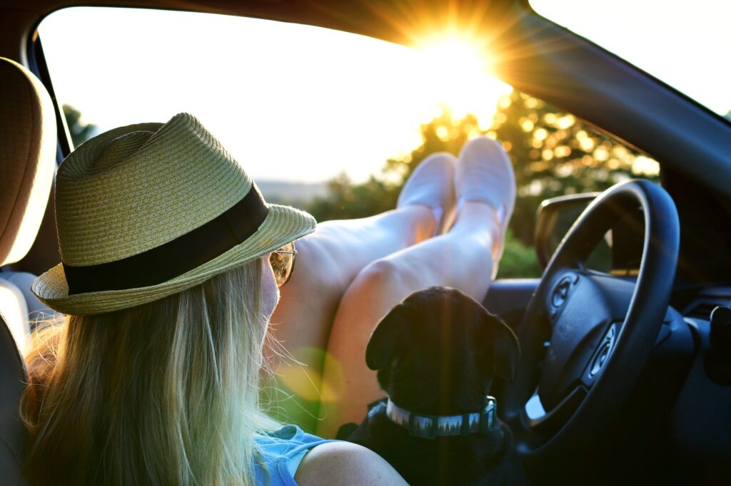 Woman with dog watching summer sunset from her car on road trip during golden hour, feet propped up
